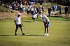 To golfspillere øver på putting på en grønn golfbane mens folk ser på i bakgrunnen.